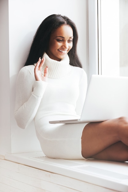 Odległość nie stanowi problemu. Szczęśliwa młoda afrykańska kobieta w ciepłym swetrze, patrząca na laptopa i machająca ręką, siedząc na parapecie