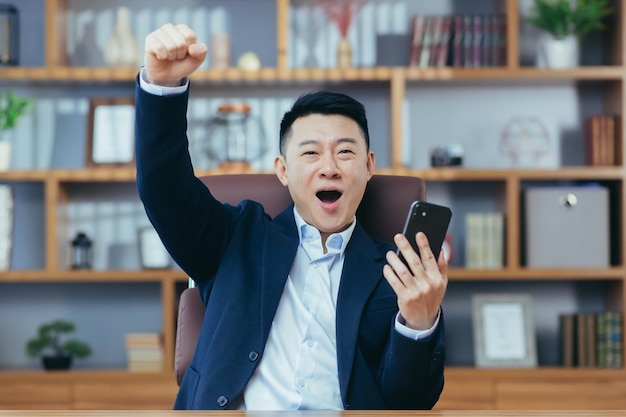 Odnosząca sukcesy Azjatka pracująca w klasycznym biurze trzymająca telefon świętuje gest zwycięstwa