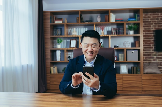 Odnoszący sukcesy azjatycki biznesmen pracujący w biurze, siedzący przy stole, piszący przez telefon, szczęśliwy i uśmiechnięty
