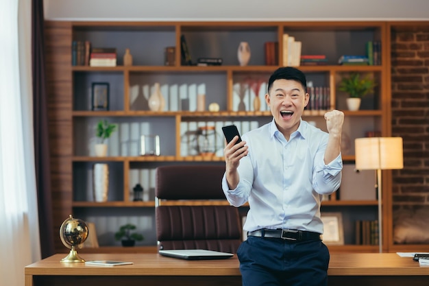 Odnoszący sukcesy azjatycki biznesmen świętuje zwycięstwo i człowiek sukcesu patrzy w kamerę i cieszy się, że wygrana telefon działa w klasycznym biurze domowym