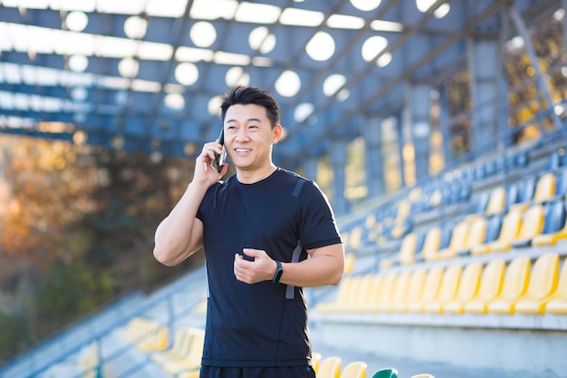 Odnoszący sukcesy azjatycki sportowiec rozmawia przez telefon, dzieląc się swoimi sportowymi osiągnięciami i wynikami, uśmiechając się i radując się na zewnątrz na stadionie sportowym