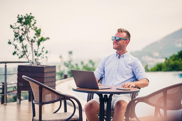 Odnoszący sukcesy przystojny biznesmen w okularach przeciwsłonecznych pracuje przy laptopie siedzącym przy basenie Praca zdalna Freelancer