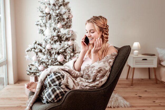 Odpoczynek w fotelu. Zadowolona jasnowłosa kobieta przyjemnie rozmawia na smartfonie podczas świąt Bożego Narodzenia w domu