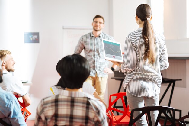 Zdjęcie odpowiadanie za pomocą laptopa. sprytna, ciekawa młoda dziewczyna stojąca z nowoczesnym laptopem w dłoniach, patrząc na mówcę podczas warsztatów rozwoju osobistego