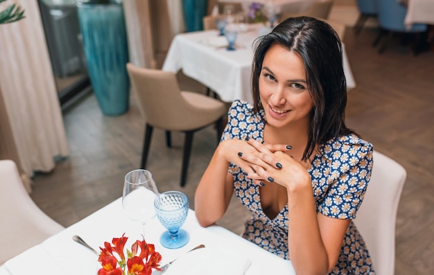 Ogólny widok uśmiechniętej młodej eleganckiej kobiety w sukience siedzącej w restauracji patrząc w kamerę Kaukaska kobieta pozuje w stołówce i czeka na swoją rodzinę na obiad Skopiuj miejsce na tekst