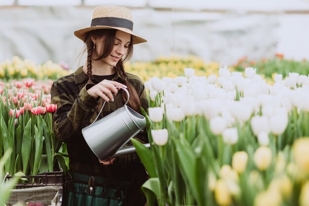Ogrodniczka w kapeluszu i rękawiczkach podlewa konewką kwietnik z tulipanów