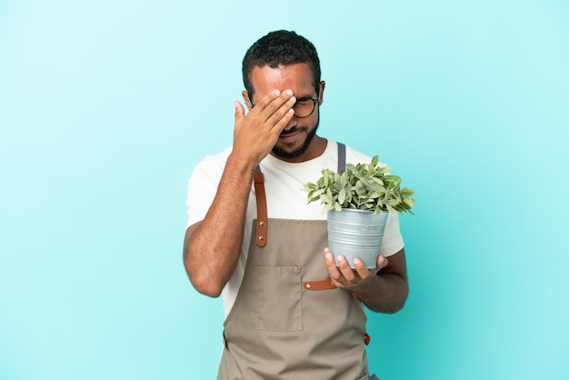 Ogrodnik latynoski mężczyzna trzymający roślinę odizolowaną na niebieskim tle ze zmęczonym i chorym wyrazem