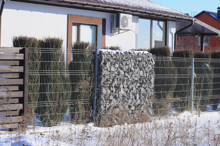 Zdjęcie ogrodzenie domu wykonane jest z kolejnych odcinków desek gabionowych oraz tui
