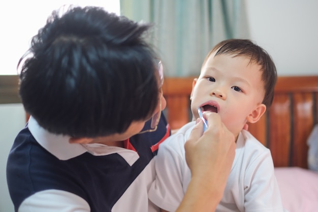 Ojciec azjatycki uczy szczotkowanie zębów dziecka, Śliczne małe dziecko w wieku 2-3 lat uczy malować zęby rano w łóżku w domu, Pielęgnacja zębów dla dzieci, Koncepcja rozwoju dziecka