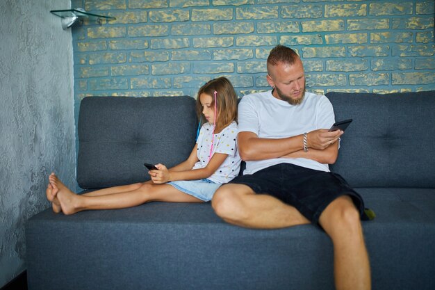 Ojciec i córka przeglądają telefon komórkowy, relaksując się razem na kanapie w domu, kobieta i dziewczyna grają na smartfonach, odpoczywając na kanapie w weekendowy dzień.