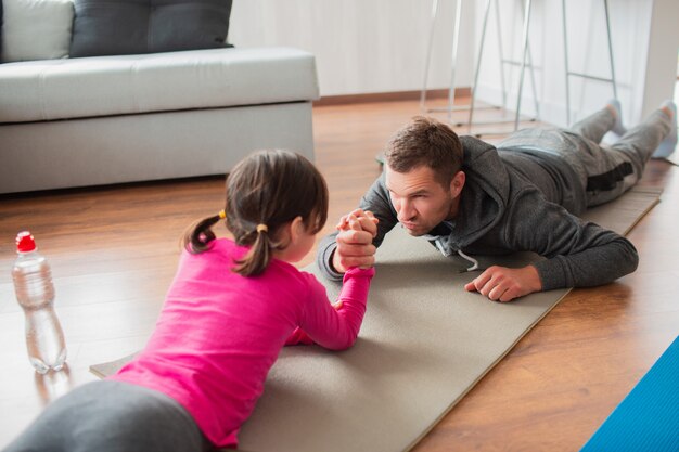 Ojciec i córka trenują w domu. Trening w mieszkaniu. Sport w domu. Weź udział w siłowaniu się na rękę i połóż się na macie do jogi