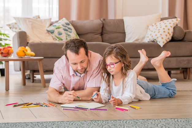 Ojciec i córka w okularach rysują razem leżąc na podłodze w pokoju mieszkalnym.