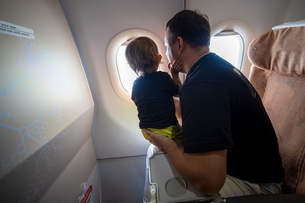 ojciec i ładny maluch siedzi w samolocie i patrząc w niebo przez iluminator. koncepcja pierwszego lotu, podróżowanie z dziećmi