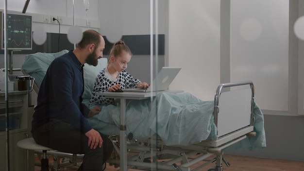 Zdjęcie ojciec siedzi z chorą córką na oddziale szpitalnym podczas grania w gry wideo terapii online przy użyciu komputera przenośnego podczas badania rekonwalescencji. dziewczyna pacjentka z rurką nosową z tlenem relaksuje się po operacji