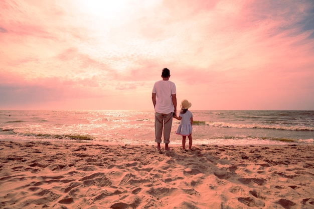 Ojciec spaceruje ze swoją córeczką po plaży wieczorem, patrząc na zachód słońca
