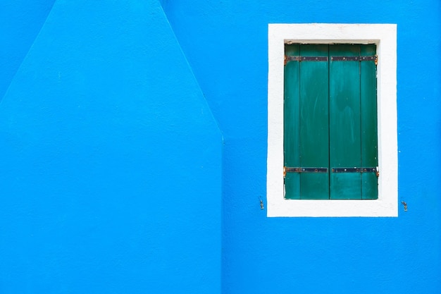 Okno z zielonymi okiennicami na niebieskiej ścianie. Kolorowa architektura na wyspie Burano, Wenecja, Włochy.