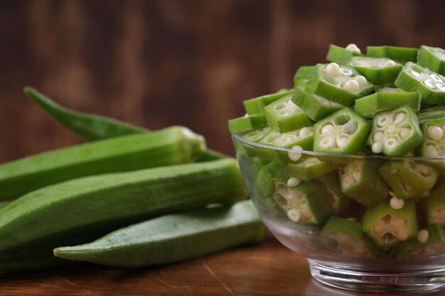 Okra lub Ladys palec lub świeże zielone warzywa Bhindi ułożone na drewnianej desce ze szklaną miską pełną pokrojonych w plasterki pierścieni okry z drewnianym tłem selektywnej ostrości