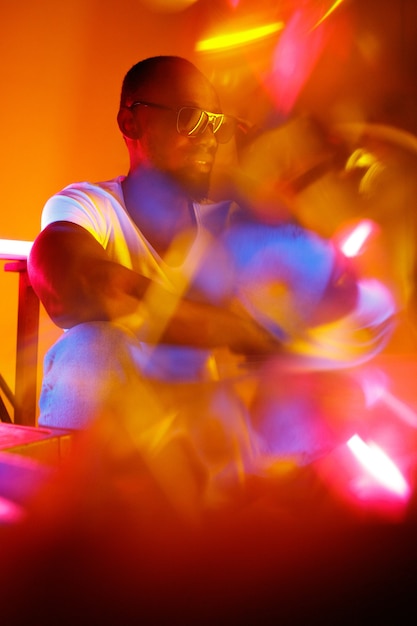 Okrutny. Kinowy portret stylowego młodego człowieka w oświetlonym neonami pokoju. Jasne, neonowe kolory. Afro-modelka, muzyk w pomieszczeniu. Kultura młodzieżowa w stylu imprezowym, festiwalowym i koncepcji muzycznej.
