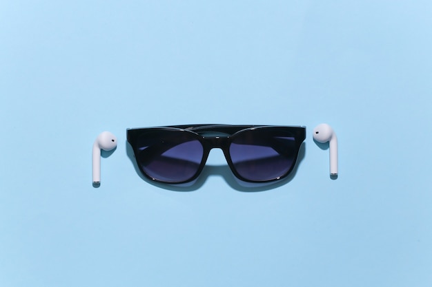 Okulary przeciwsłoneczne i białe prawdziwe bezprzewodowe słuchawki bluetooth na jasnym niebieskim tle.
