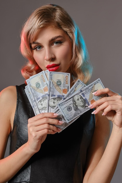 Olśniewająca blondynka dziewica z jasnym makijażem w czarnej stylowej sukience trzyma trochę pieniędzy i pozuje na szarym tle studia Hazard rozrywka poker kasyno Zbliżenie