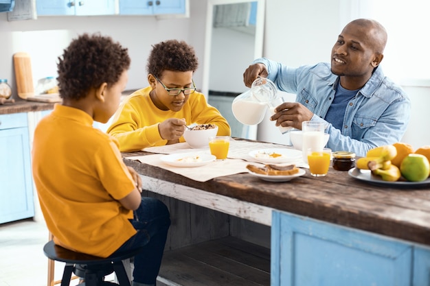 Omawianie planów. Optymistyczny młody człowiek nalewa szklankę mleka i rozmawia z synami, kiedy wszyscy jedzą śniadanie w kuchni