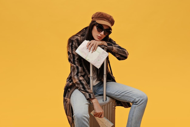 Opalona dama w stylowym płaszczu okularach przeciwsłonecznych i berecie trzyma bilety na mapę i siedzi na walizce na żółtym tle