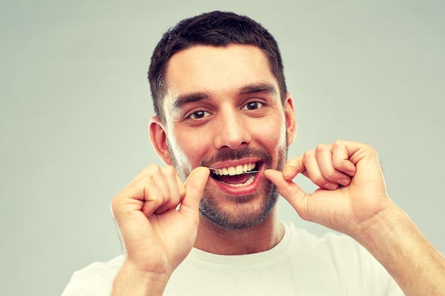 opieka zdrowotna, higiena zębów, ludzie i koncepcja piękna - uśmiechnięty młody człowiek z nicią dentystyczną czyści zęby na szarym tle