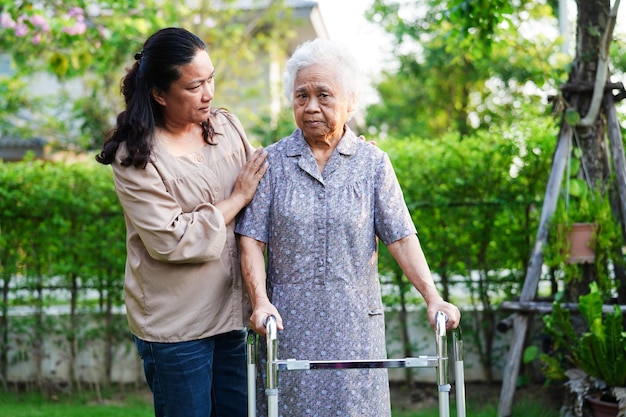 Opiekun pomaga azjatyckiej starszej kobiety z niepełnosprawnością chodzącą z chodzikiem w koncepcji medycznej w parku