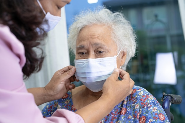 Opiekun pomaga azjatyckiej starszej lub starszej starszej kobiecie siedzącej na wózku inwalidzkim i noszącej maskę na twarz w celu ochrony przed infekcją Covid 19 Koronawirus