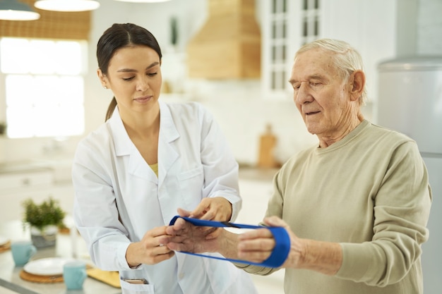 Opiekuńcza pielęgniarka w białym fartuchu wyjaśniająca emerytowi, jak wykonywać ćwiczenia w domu z gumką fitness