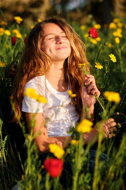 Optymistyczna dziewczyna uśmiechająca się i wąchająca świeże kwiaty z zamkniętymi oczami podczas relaksu na trawiastym polu