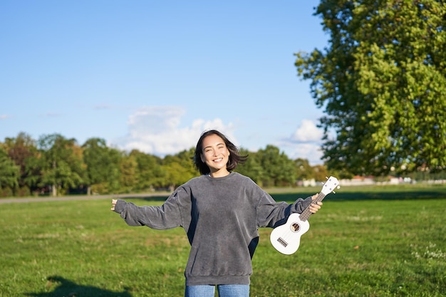 Optymistyczna młoda kobieta tańcząca ze swoją dziewczyną na instrumencie muzycznym podnosi ukulele i pozuje w parku o