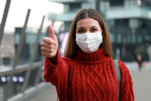 Optymistyczna młoda kobieta ubrana w maskę ochronną pokazując kciuki do góry