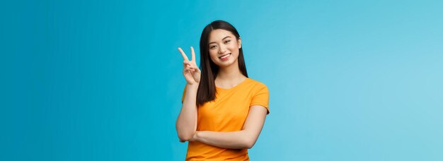 Optymistyczna słodka, delikatna azjatycka młoda tysiącletnia dziewczyna wysyła pozytywne wibracje, przechyla głowę i pokazuje pokojowe zwycięstwo