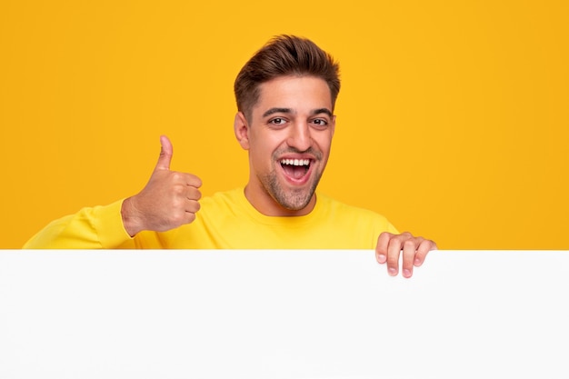 Optymistyczny Młody Brodaty Facet Uśmiecha Się Radośnie I Pokazuje Aprobujący Gest Kciuka, Stojąc Za Pustym Białym Plakatem Na żółtym Tle