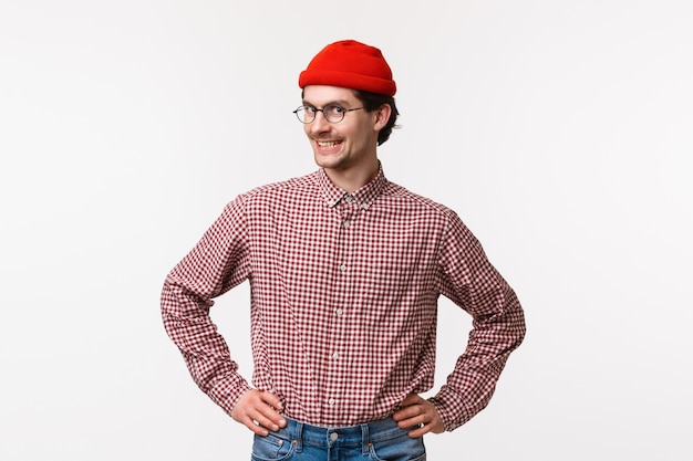 Optymistyczny portret w pasie i entuzjastyczny młody hipster w czerwonej czapce i okularach, w kraciastą koszulę, stojący z pewną, prostą pozą, trzymający ręce w pasie i uśmiechnięty aparat