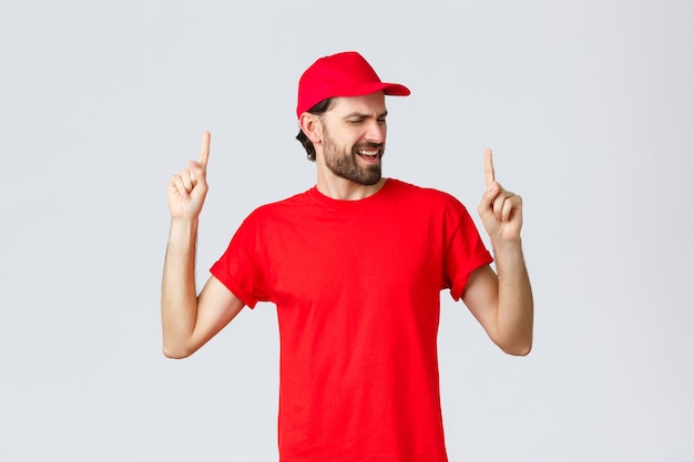 Optymistyczny przystojny brodaty kurier spędza przerwę na relaks pracownika w czerwonej czapce mundurowej i koszulce ...