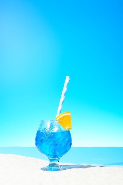 Orzeźwiający niebieski napój z lodem i plasterkiem pomarańczy w szklance na piaszczystym wybrzeżu