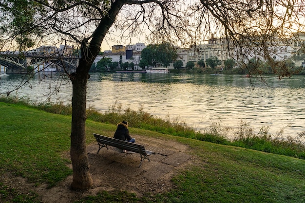 Osoba siedząca na ławce w publicznym parku, patrząc na rzekę, gdy słońce zachodzi za budynkami. Hiszpania.