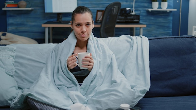 Osoba z grypą sezonową owinięta kocem na kanapie. Chora kobieta czuje zimno, trzymając filiżankę herbaty przeciwko chorobie i patrząc w kamerę. Dorosły z napojem i lekarstwami na chorobę