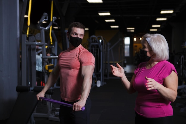 Osobisty trener i jego starsza klientka nosząca medyczne maski na twarz podczas treningu na siłowni