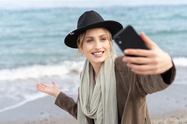 Oszałamiająca młoda kobieta robi selfie i pokazuje piękną plażę z wielkim uśmiechem za pomocą telefonu komórkowego