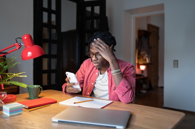 Oszołomiony, Zdezorientowany Afroamerykanin Czyta Wiadomości W Telefonie Komórkowym, Siedząc Przy Biurku Z Laptopem