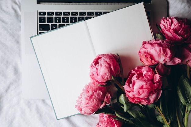 Otwarty notebook, laptop i różowe kwiaty peonii na białej pościeli.