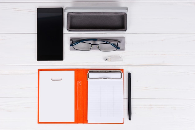 Otwórz pamiętnik z długopisem do organizowania harmonogramu, okulary i otwórz etui na okulary w pobliżu telefonu komórkowego na drewnianym białym stole