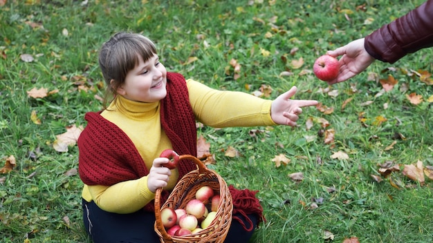 Zdjęcie otyła dziewczyna w czerwonym szaliku siedzi na trawie i jesienią trzyma kosz z jabłkami