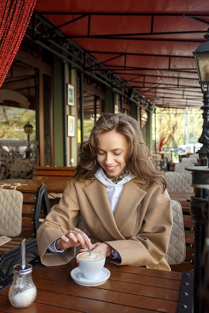 Outdoors portret młoda uśmiechnięta dziewczyna jest ubranym beżowego żakiet pije kawę na cukiernianej werandzie miesza cukier z łyżką