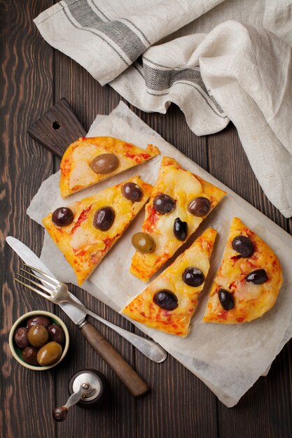 Owalna pizza grecka z oliwkami i pomidorem pokrojona na kawałki na ciemnym drewnianym rustykalnym stylu.