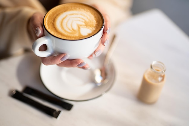 Pachnący i ciepły kubek cappuccino z rysunkowym serduszkiem trzymanym w dłoniach