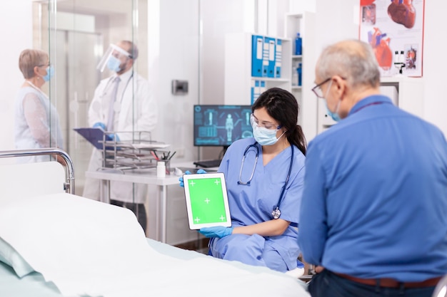 Zdjęcie pacjent patrzący na tablet z zielonym ekranem podczas konsultacji w sali szpitalnej z pielęgniarką noszącą maskę przeciw koronawirusowi jako środek ostrożności